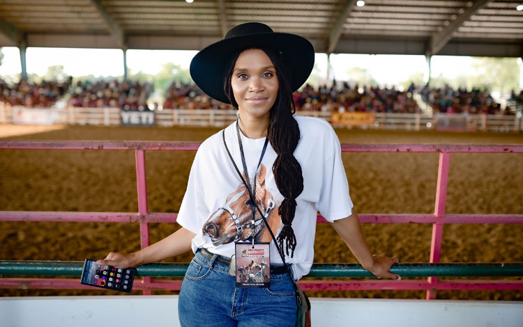 A woman wearing a hat looking directly at the camera, holding a cell phone and posing.